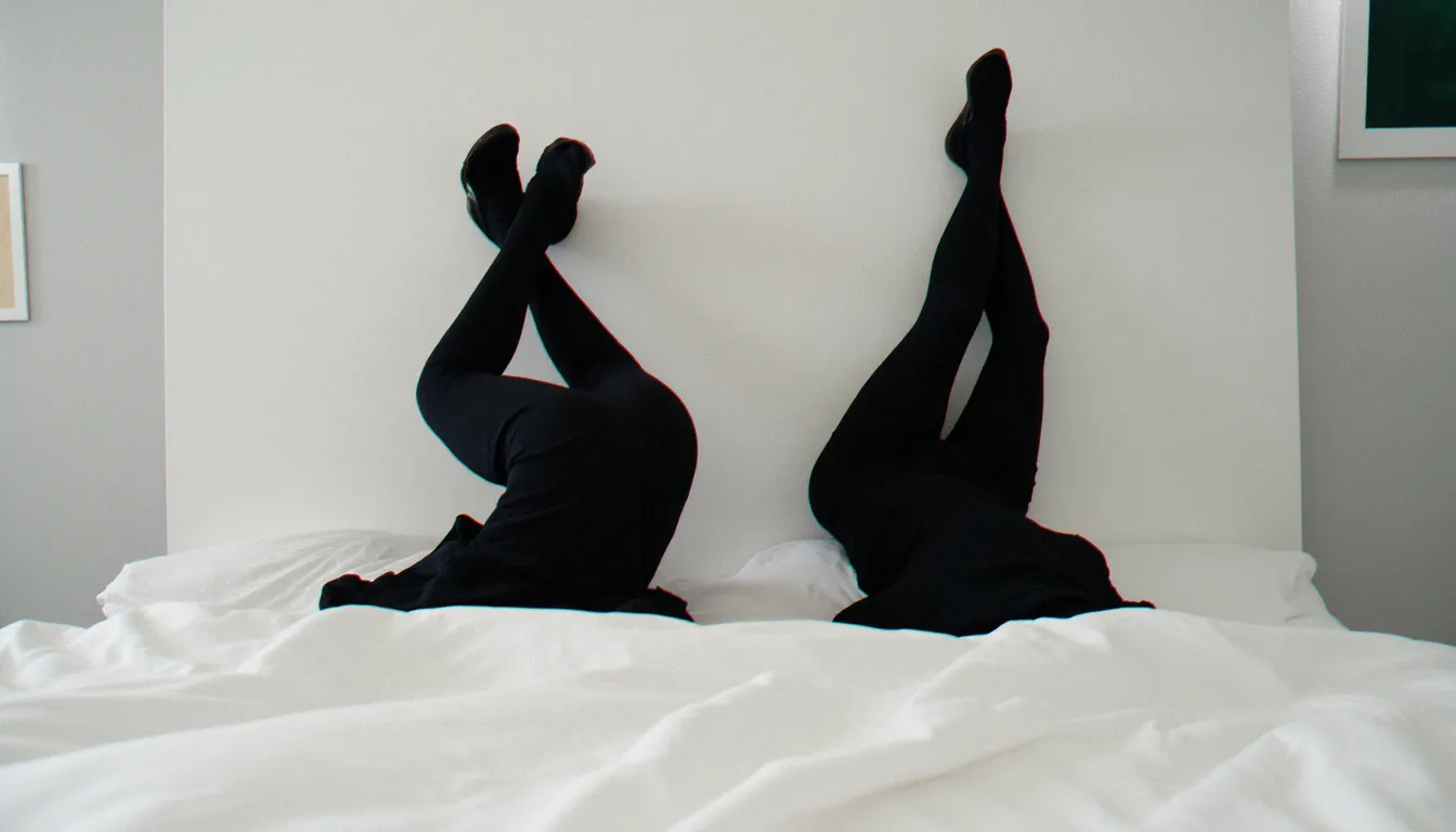 Silhouettes de deux partenaires allongés tête-bêche sur un lit blanc minimaliste, illustrant la position 99 dans un cadre sobre et artistique